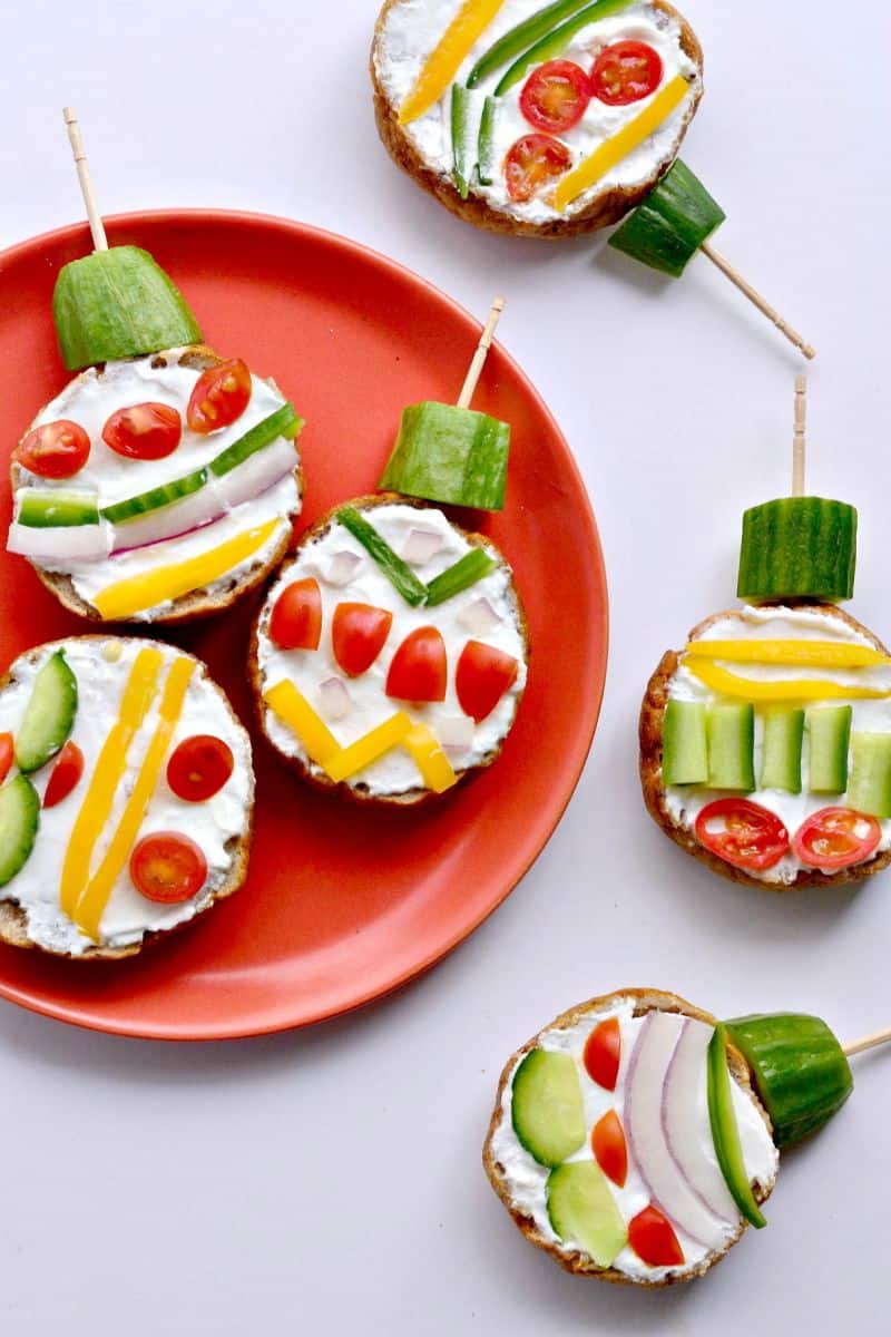 bagels cut in half slathered with white cream cheese and decorated to look like christmas ornaments with a variety of different veggies