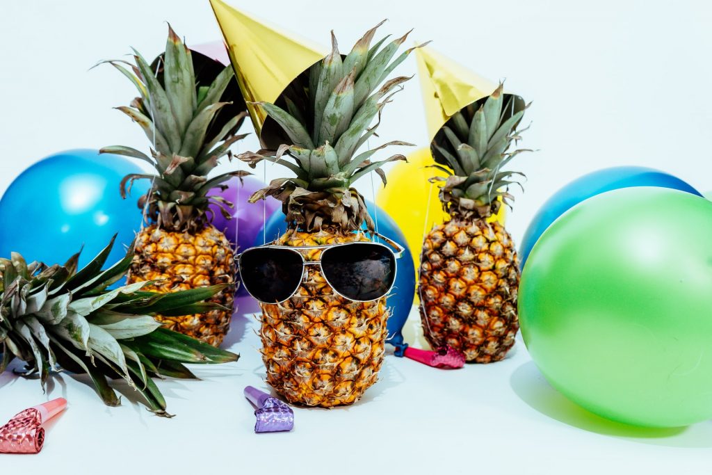 pineapples on a table with sunglasses and party hats surrounded by balloons and party favors